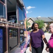 An Artman employee in a purple shirt and a resident in a pink shirt wait for frozen ice at a Kona Ice Truck.